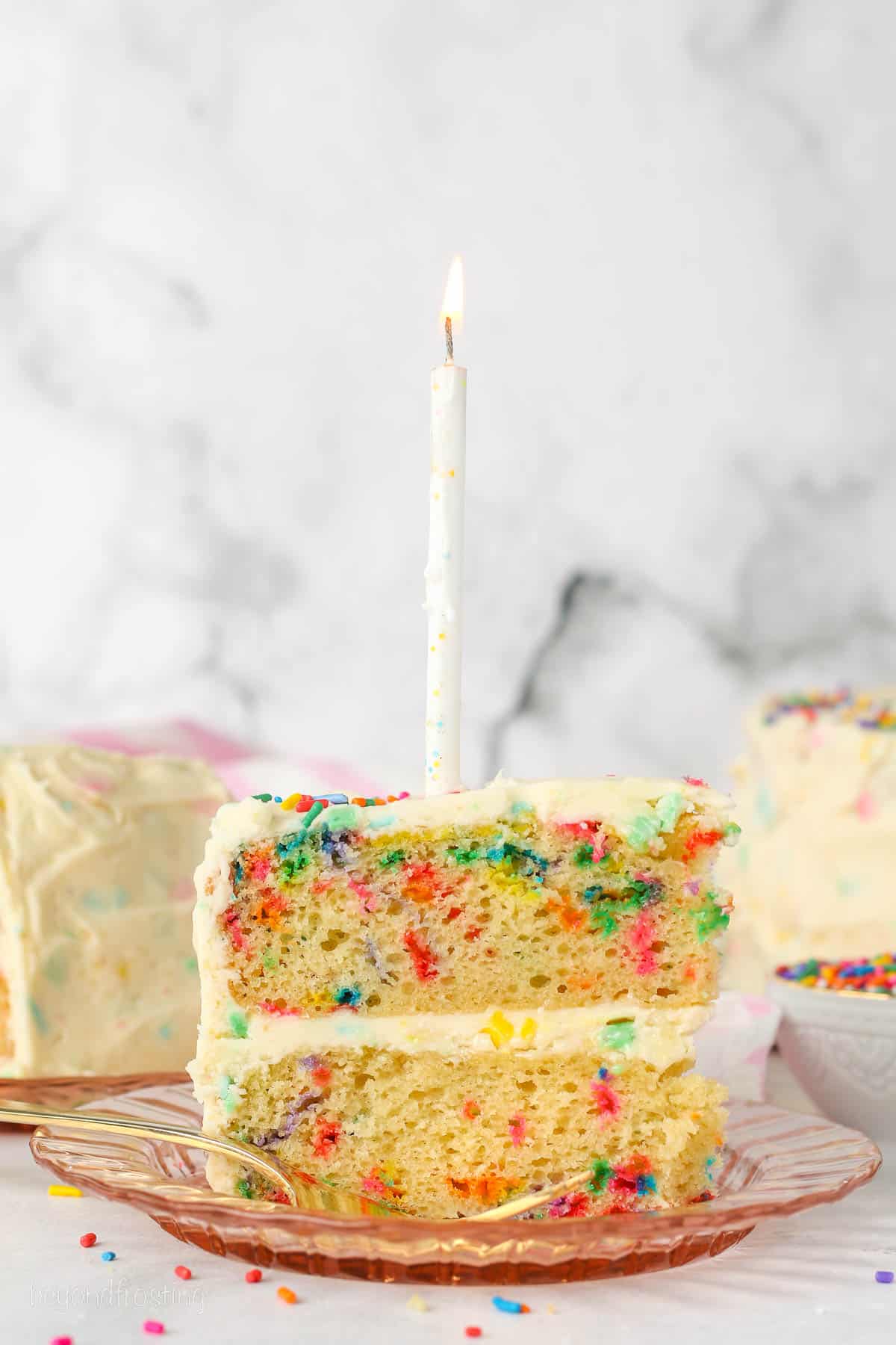 A slice of frosted confetti cake topped with a birthday candle.