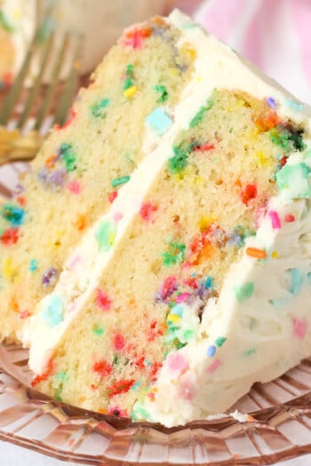 A slice of confetti cake frosted with confetti frosting on a plate.
