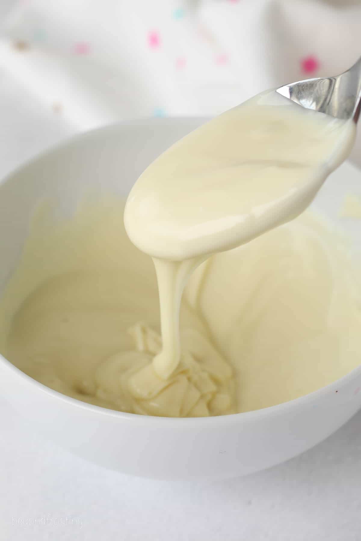A spoon drizzles melted white chocolate into a bowl.