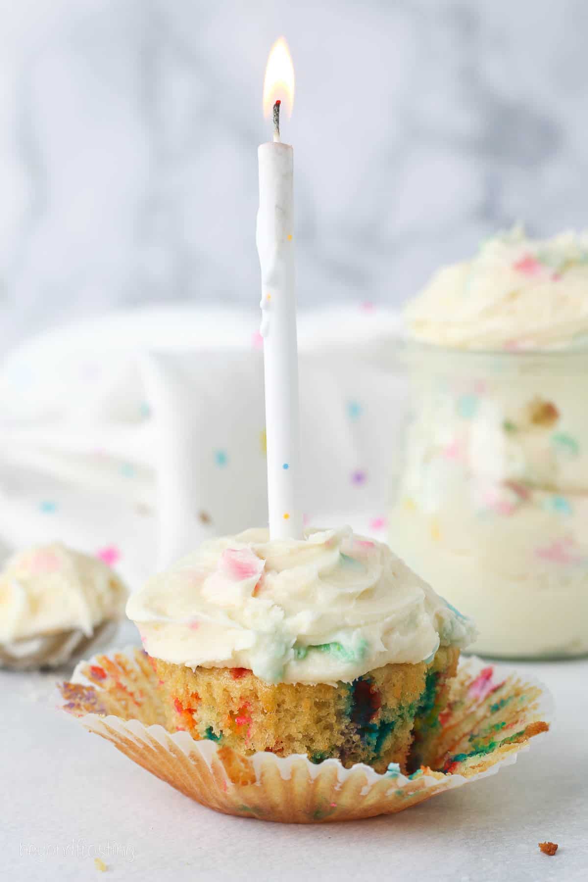 A partially unwrapped funfetti cupcake frosted with rainbow chip frosting and topped with a lit birthday candle.