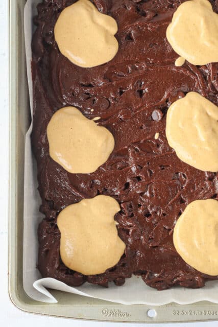 Dollops of pumpkin cheesecake batter layered over chocolate brownie batter in a parchment lined baking pan.