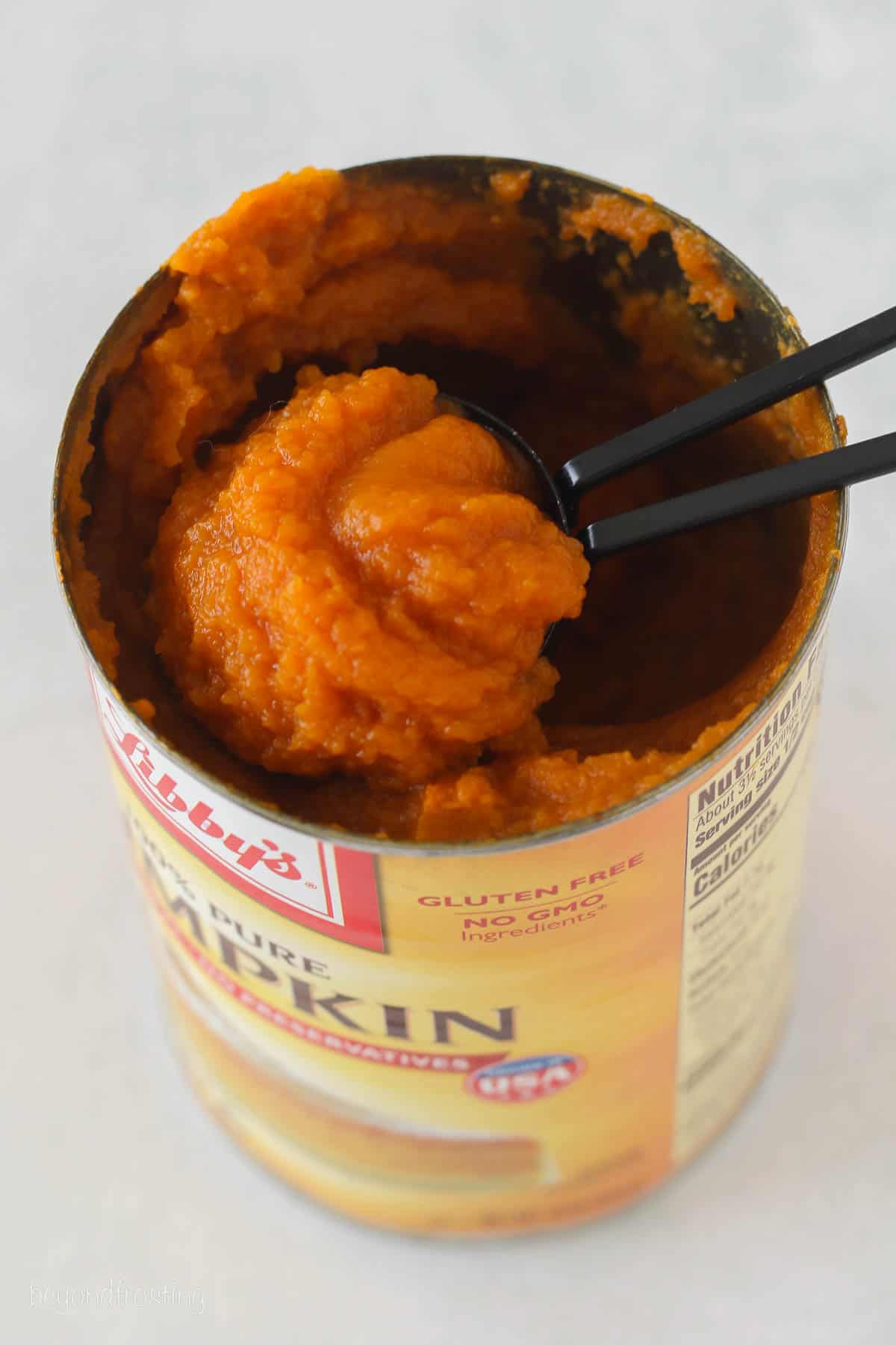 A measuring spoon scoops pumpkin from an open can of pumpkin puree.