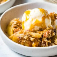 A serving of apple crisp in a white bowl topped with a scoop of ice cream and drizzled with caramel sauce.