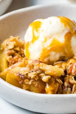 A serving of apple crisp in a white bowl topped with a scoop of ice cream and drizzled with caramel sauce.