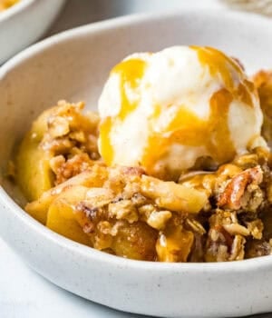 A serving of apple crisp in a white bowl topped with a scoop of ice cream and drizzled with caramel sauce.