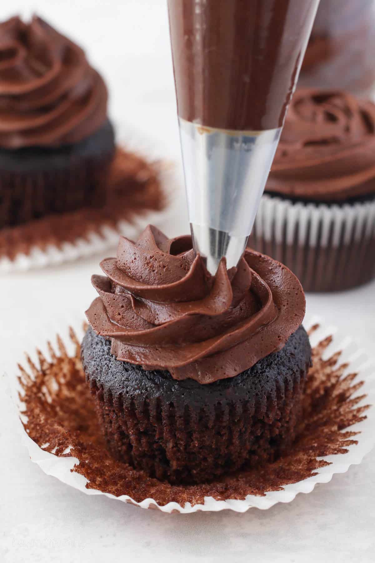 A piping tip pipes a swirl of chocolate ganache frosting onto a chocolate cupcake, with more frosted cupcakes in the background.