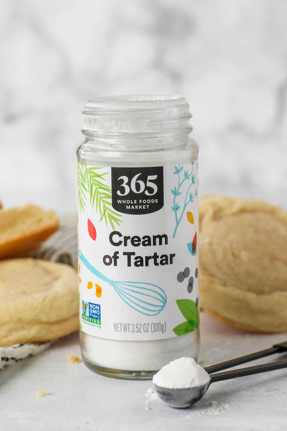 A jar of cream of tartar on a countertop next to a teaspoonful, with sugar cookies in the background.