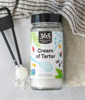 Overhead view of a jar of cream of tartar laying on a countertop next to a teaspoonful.
