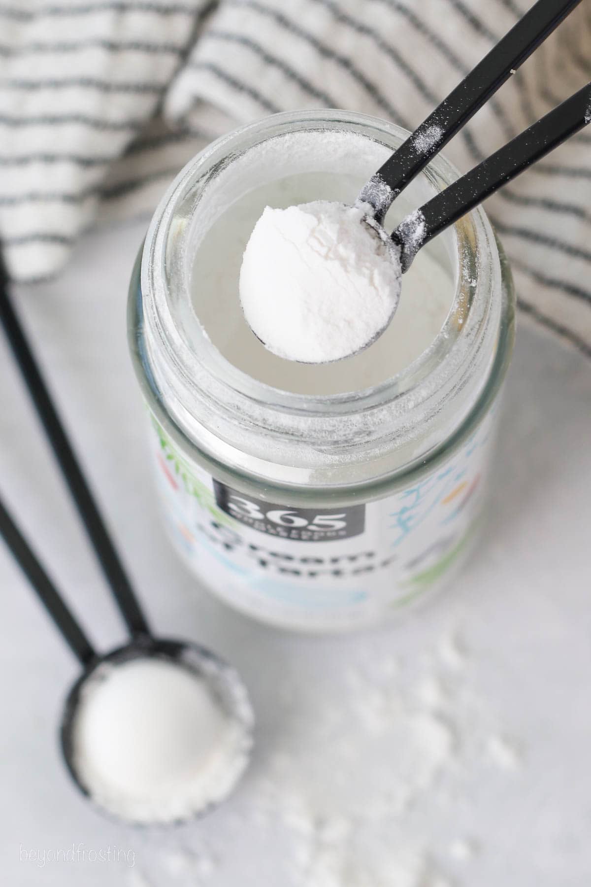 A teaspoon of cream of tartar held over a jar, next to a second teaspoonful on a countertop.