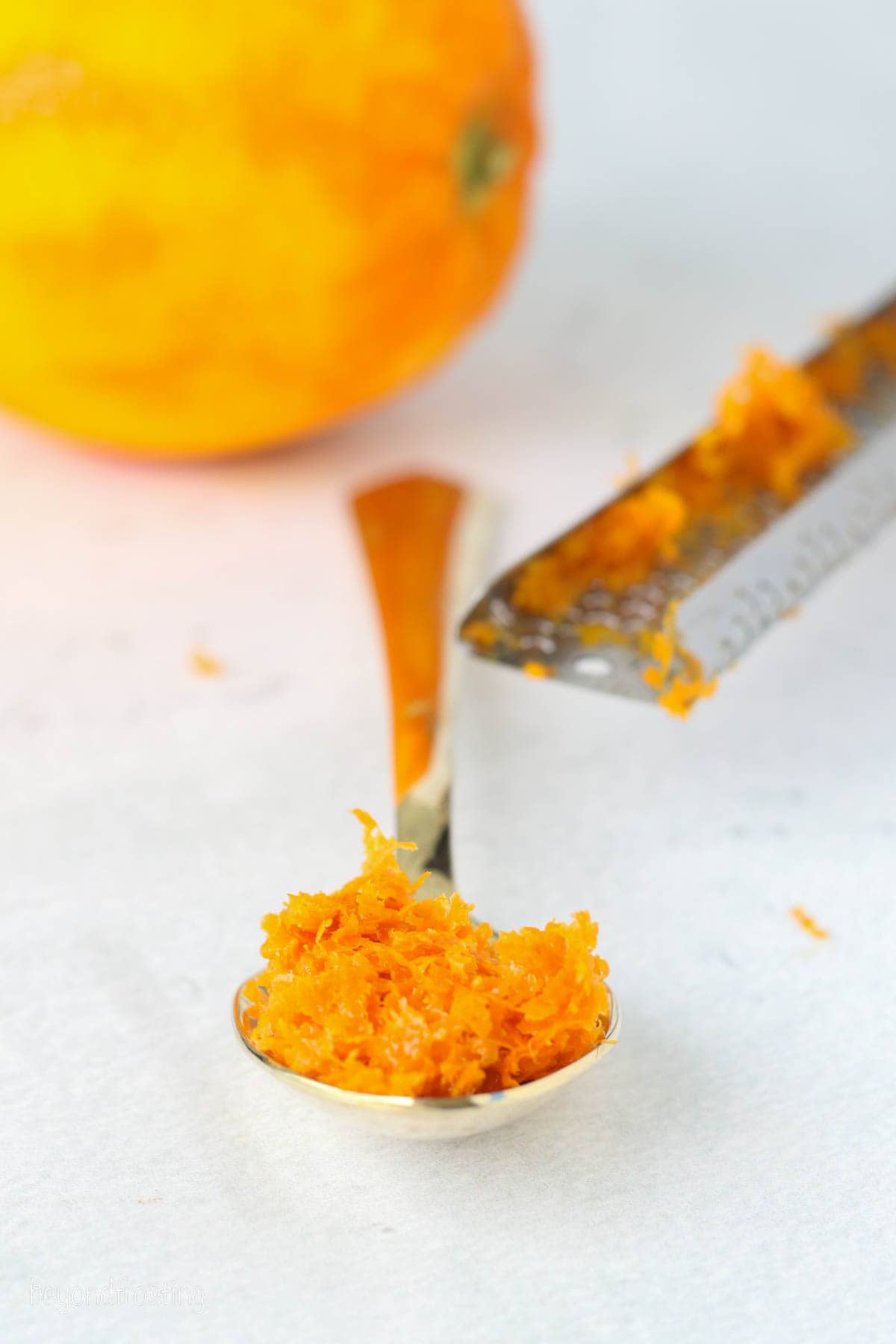 A spoonful of fresh orange zest next to a citrus zester on a countertop, with a zested orange in the background.