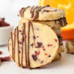 A cranberry orange shortbread cookie drizzled with chocolate leaning against a stack of cookies.