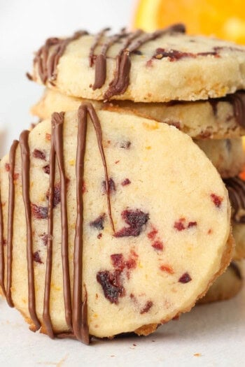 A cranberry orange shortbread cookie drizzled with chocolate leaning against a stack of cookies.