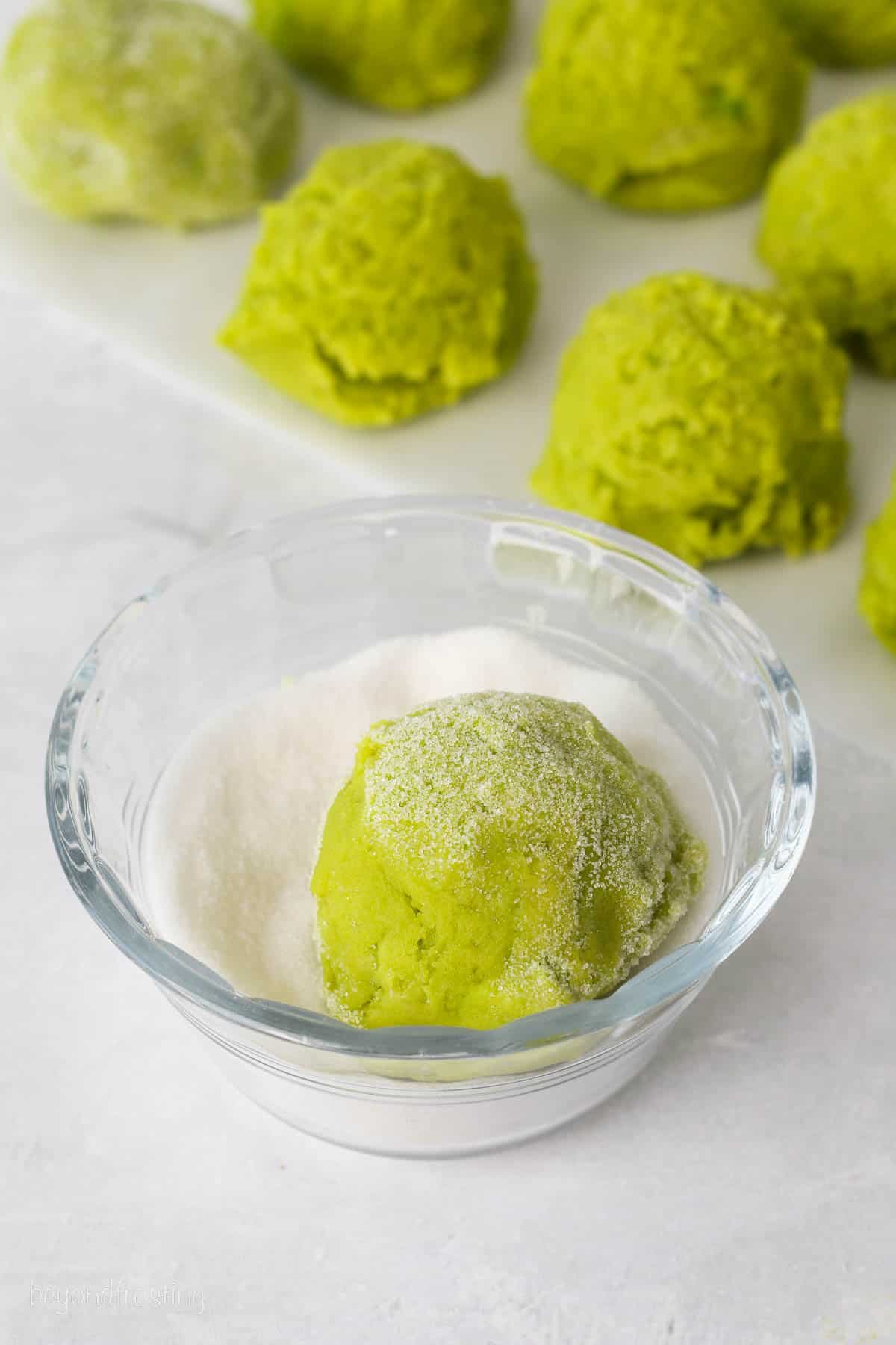 A green sugar cookie dough ball dropped in a bowl of sugar, with more green cookie dough balls in the background.