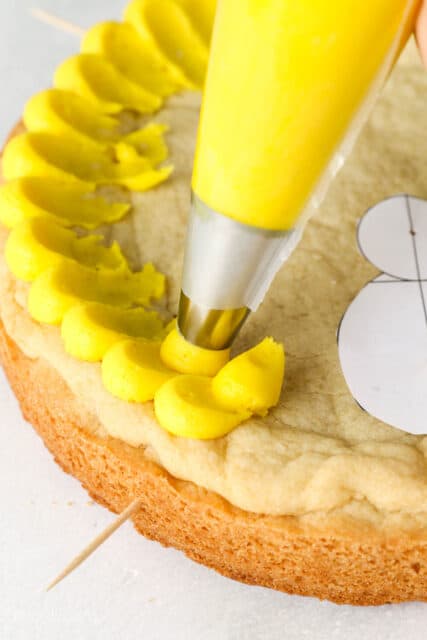Piping bag with yellow buttercream demonstrating a pedal technique on a cookie cake