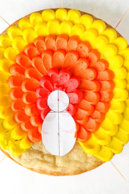 Process shot of decorating a cake with a turkey design using yellow, orange and red buttercream with a template