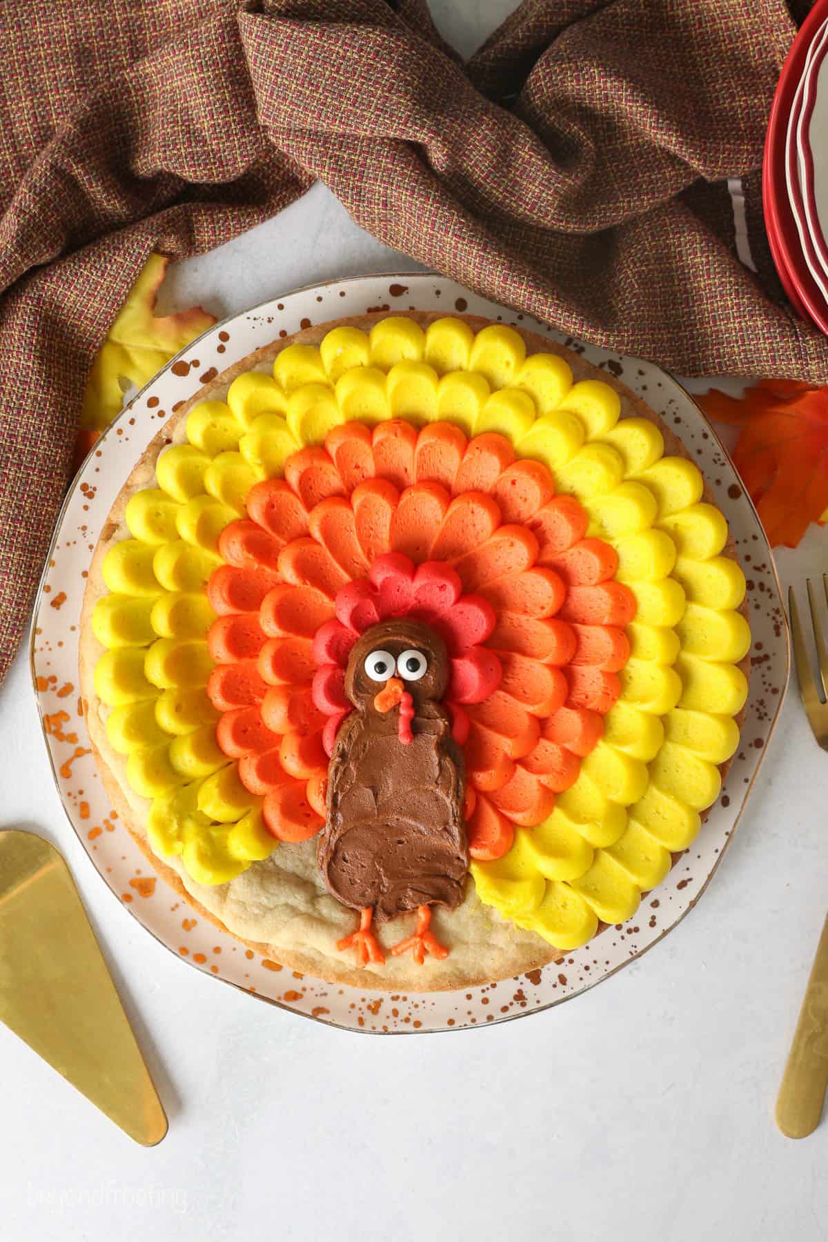 A cookie cake decorated with buttercream to look like a turkey on a plate surrounded with plates, napkin and utensils.
