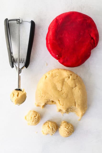 A cookie scoop next to partially portioned white cookie dough, and a disc of red cookie dough,