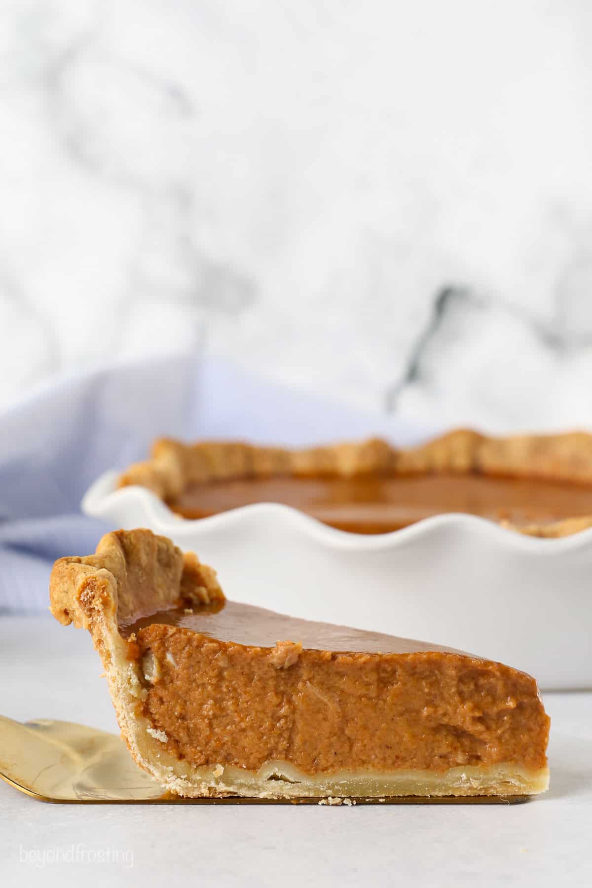 Side view of a slice of pumpkin pie in a blind baked pie crust, with the rest of the pie in the background.
