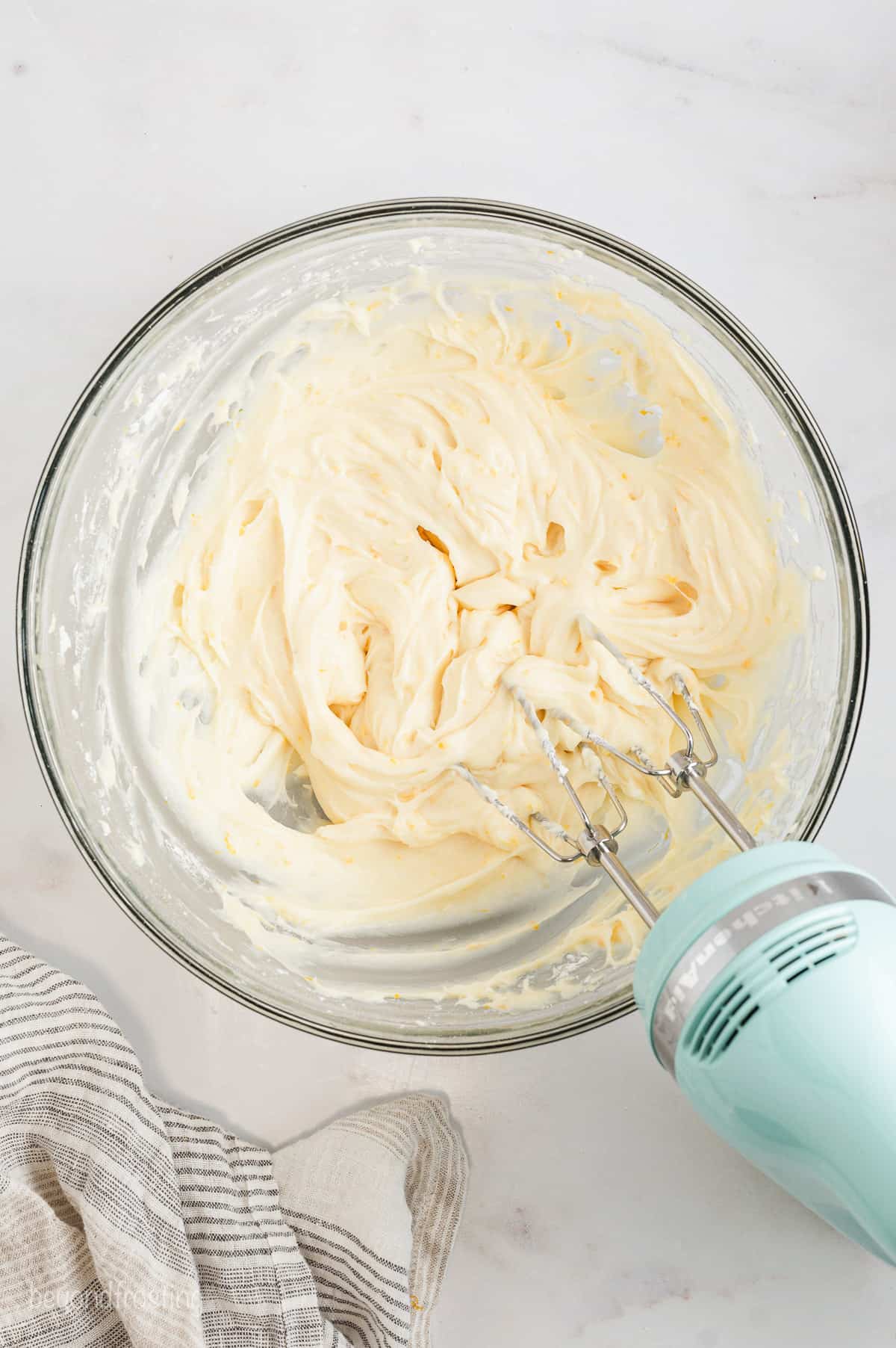 A hand mixer resting in a glass bowl of whipped orange icing.