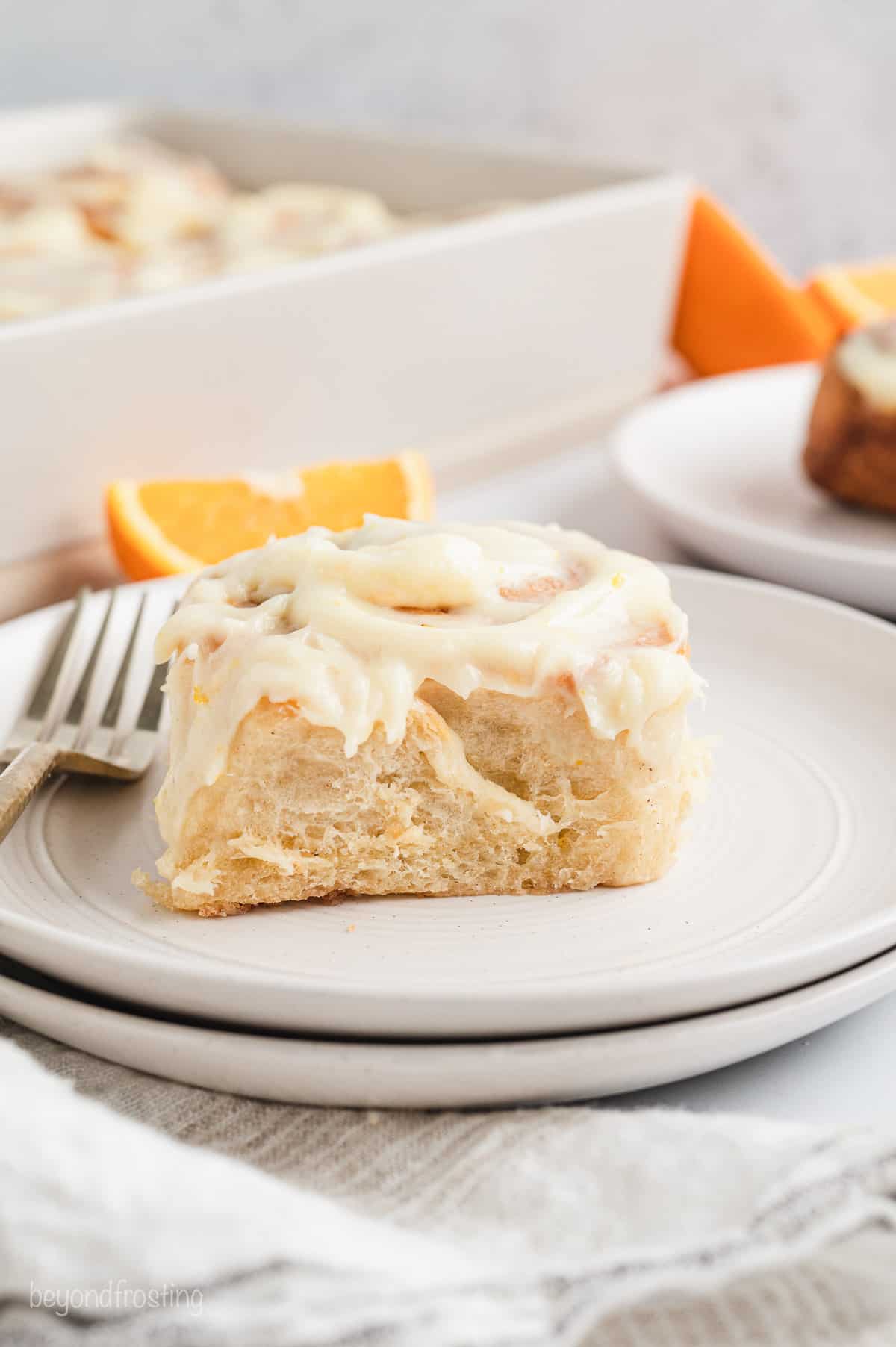 An orange roll on a white plate next to a fork, with more rolls in a baking dish in the background.