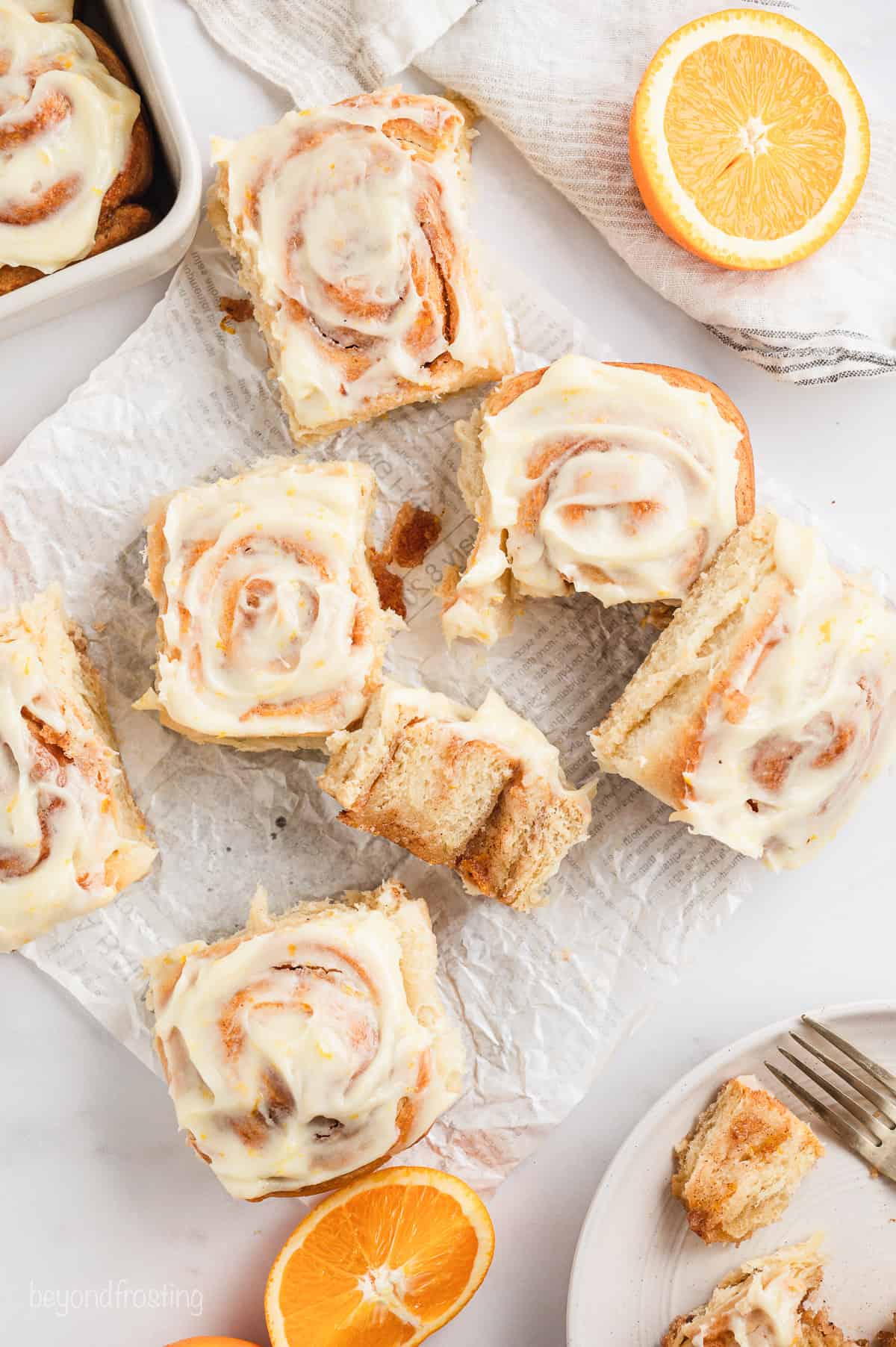 Overhead view of assorted orange cinnamon rolls on a piece of parchment paper, next to orange wedges.
