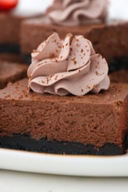 A close up of a sliced chocolate cheesecake bar on a white rimmed plate topped with chocolate whipped cream