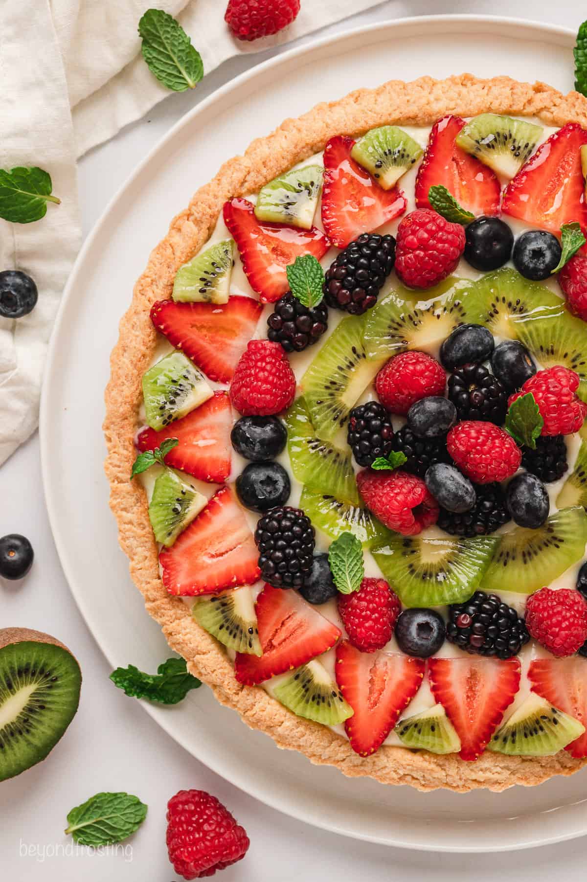 Overhead close up view of a fruit tart surrounded by scattered fresh fruit.