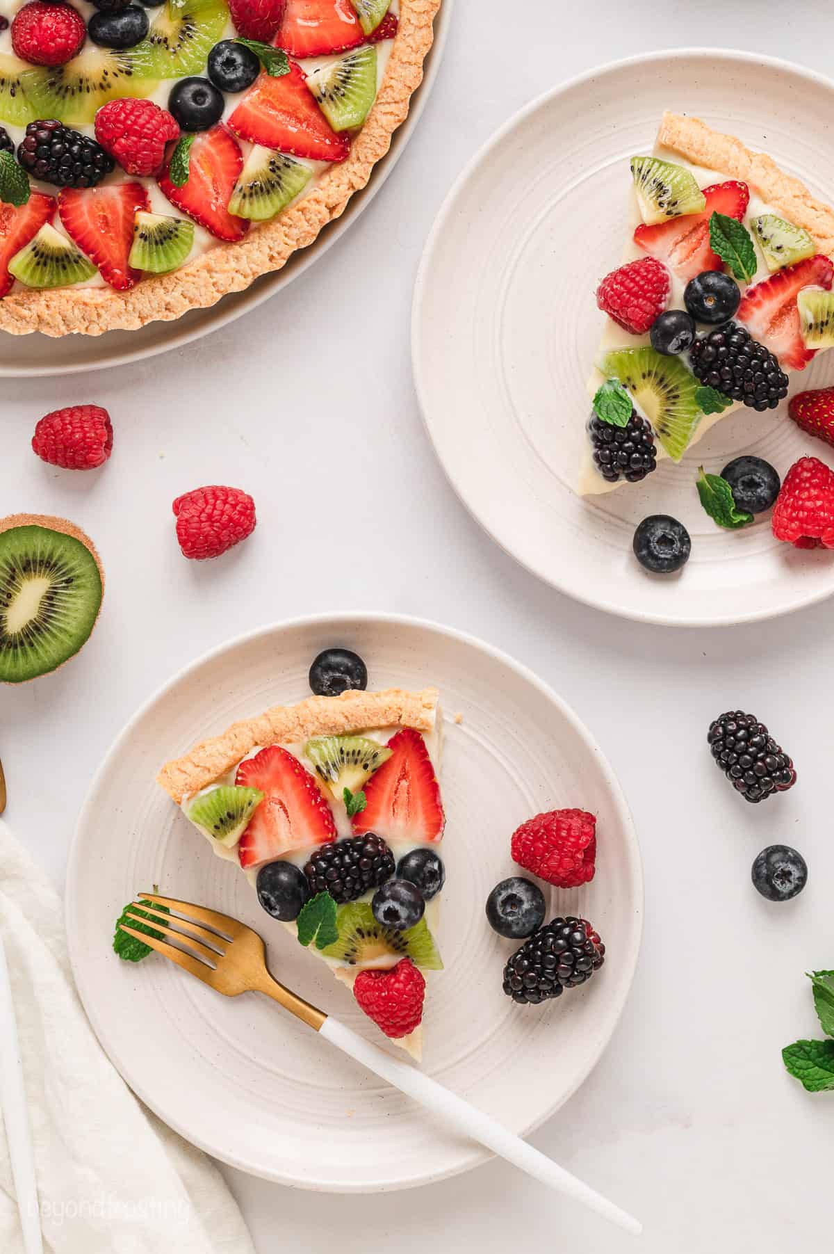 Overhead view of two slices of fruit tart served on white plates, next to the rest of the fruit tart on a countertop.