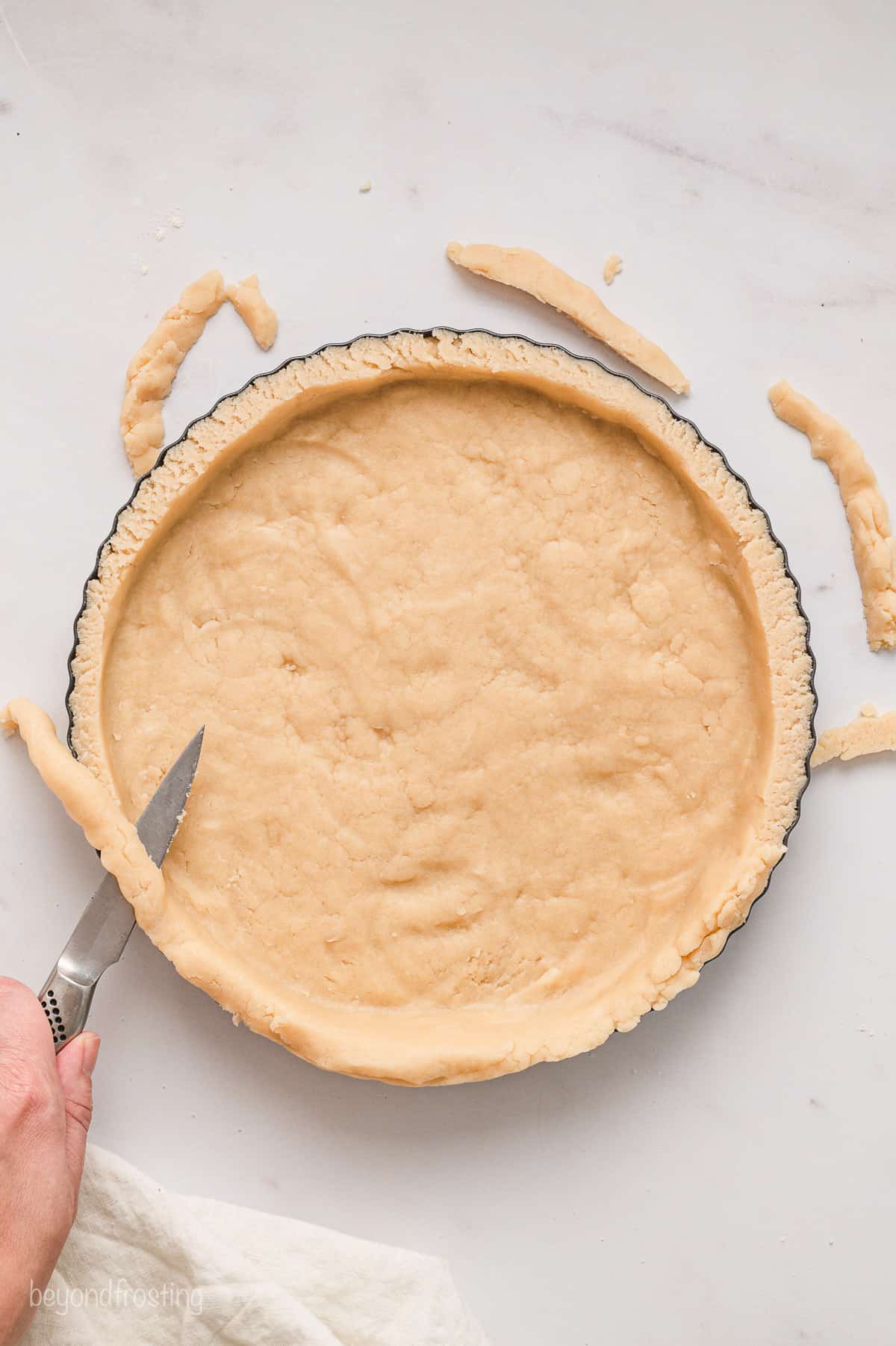 A hand uses a knife to trim away the excess pastry crust from a pie plate.