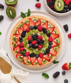 Overhead view of a fruit tart surrounded by scattered fresh fruit, next to more fruit in a bowl.