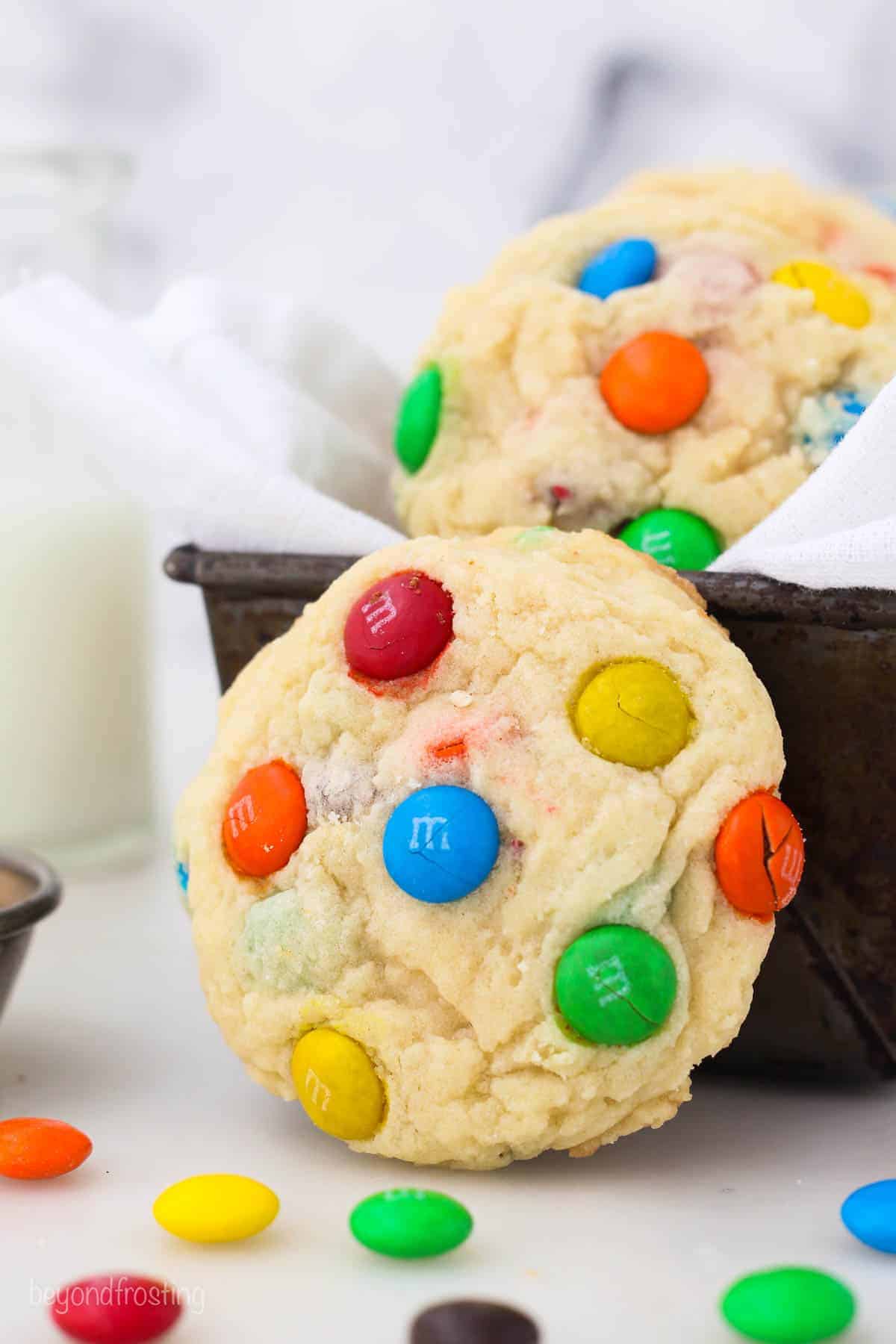 An M&M cookie propped upright against a cloth-lined tin full of more cookies, surrounded by scattered M&Ms.