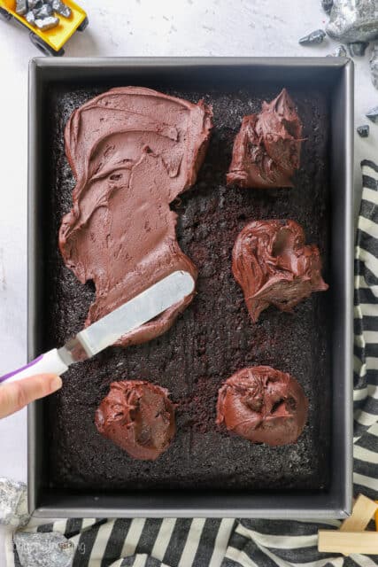 A hand uses an offset spatula to spread chocolate frosting over a 9x13-inch chocolate cake in a cake pan.