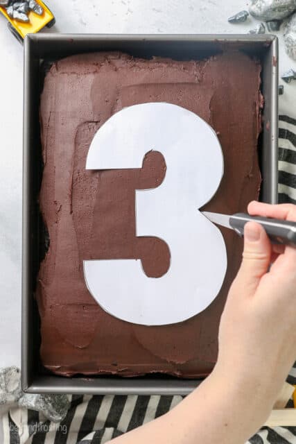 A hand uses a sharp knife to trace the outline of a paper cut-out of the number 3 on a frosted chocolate sheet cake.