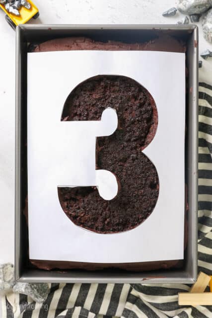 A piece of paper with the number 3 cut out, placed over a frosted chocolate sheet cake.