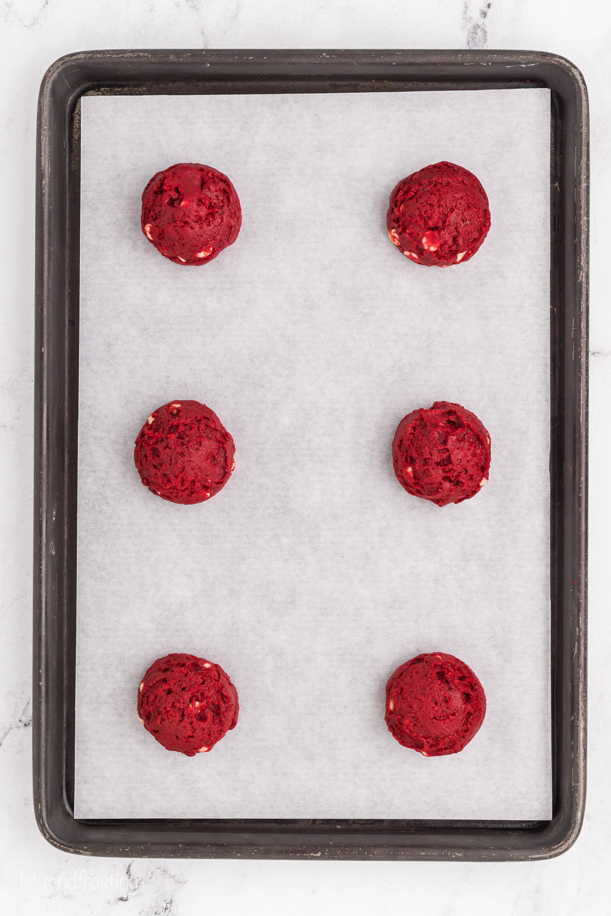 Six red velvet cookie dough balls arranged on a parchment-lined baking sheet.