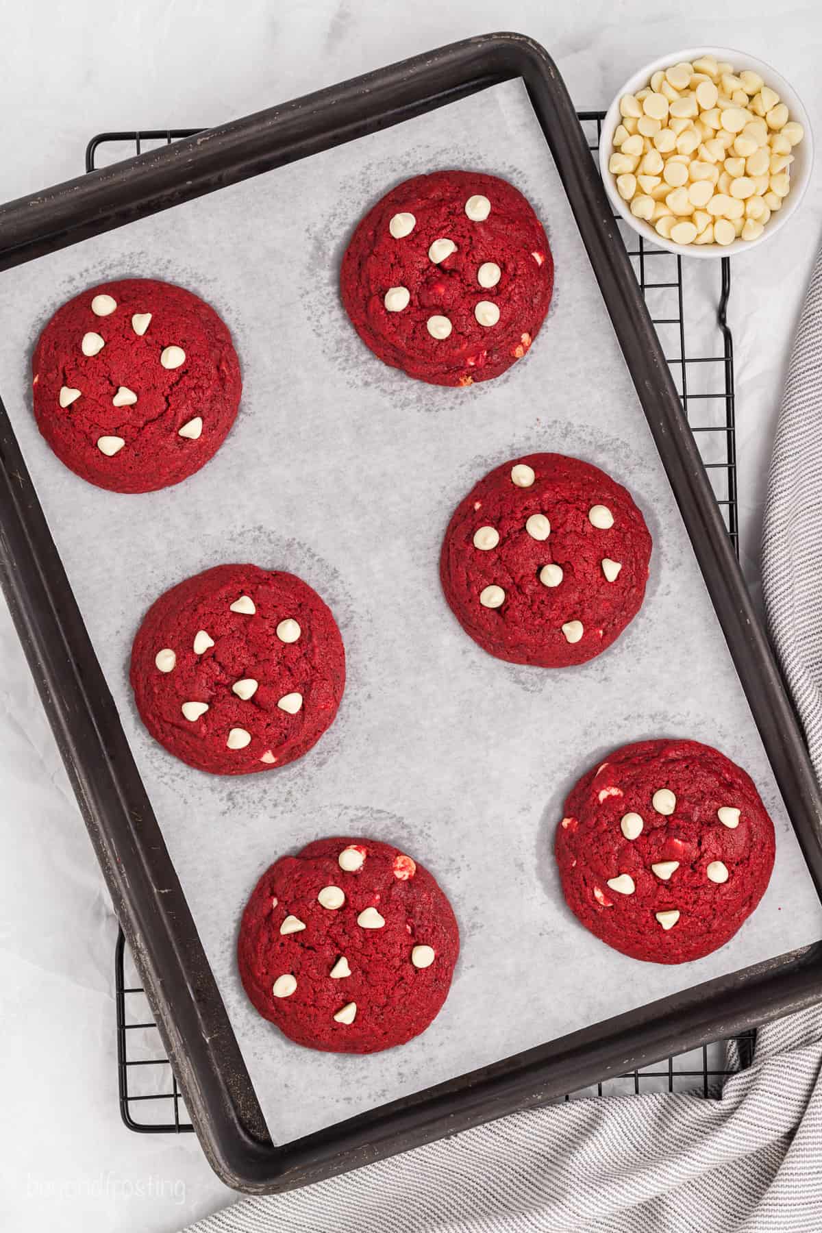 Overhead view of six red velvet cookies topped with white chocolate chips on a parchment-lined baking sheet.