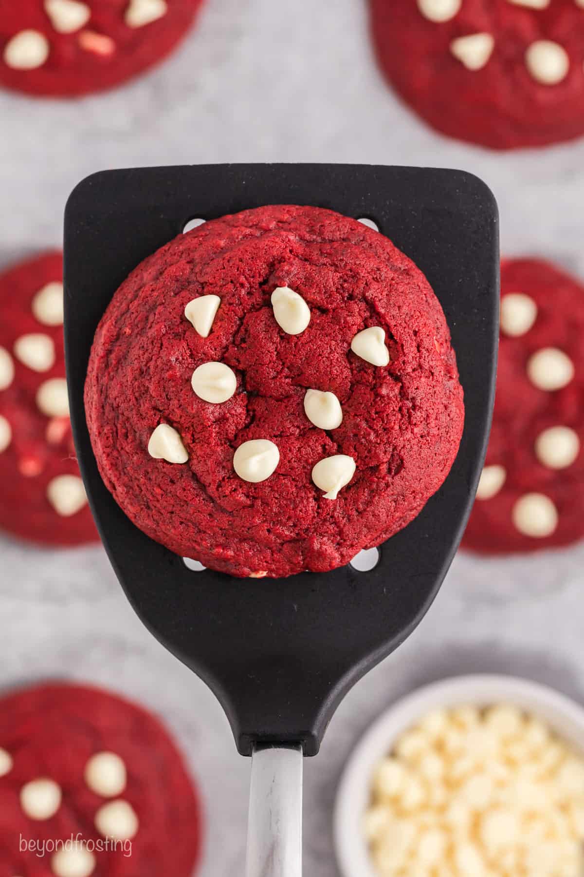 Overhead close up view of a spatula holding a red velvet chocolate chip cookie over more cookies on a baking sheet next to a bowl of white chocolate chips.