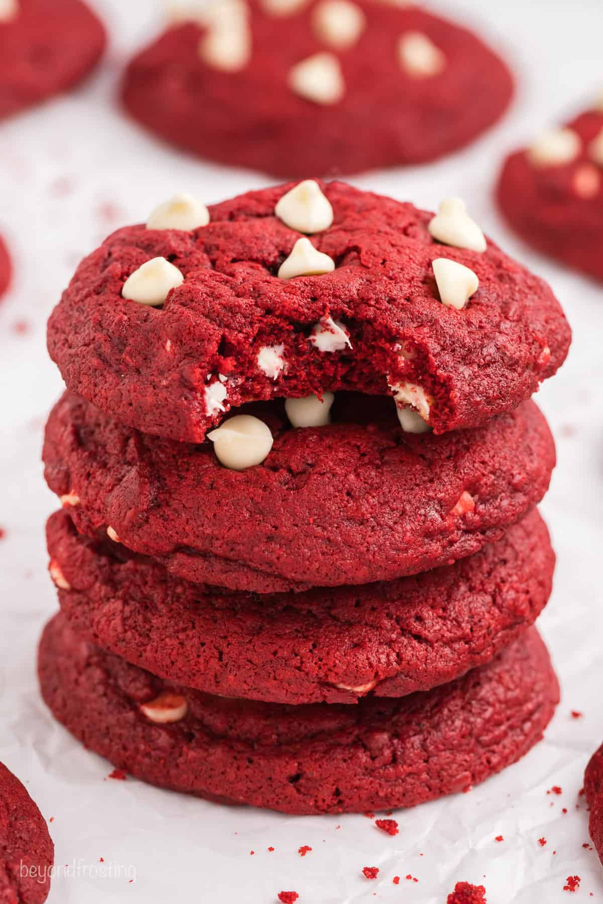 A stack of four red velvet chocolate chip cookies with a bite missing from the top cookie, and more cookies in the background.
