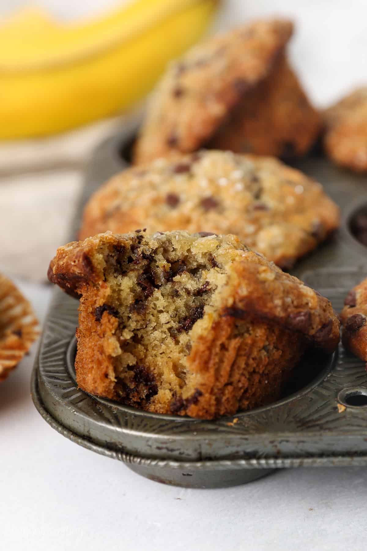 A banana chocolate chip muffin with a bite missing resting in the well of a muffin pan.