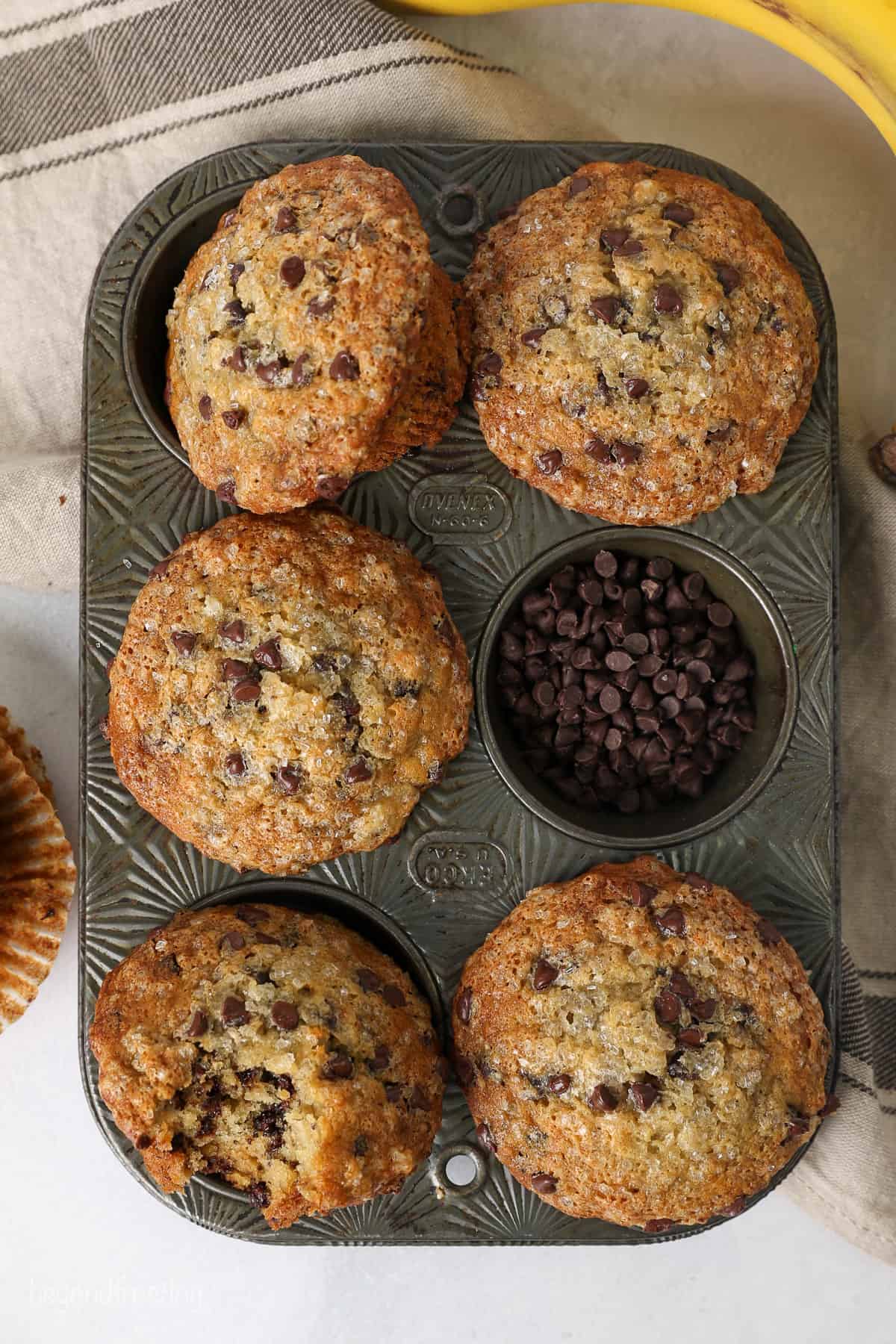 Overhead view of banana chocolate chip muffins in a muffin tin, with one well filled with chocolate chips.