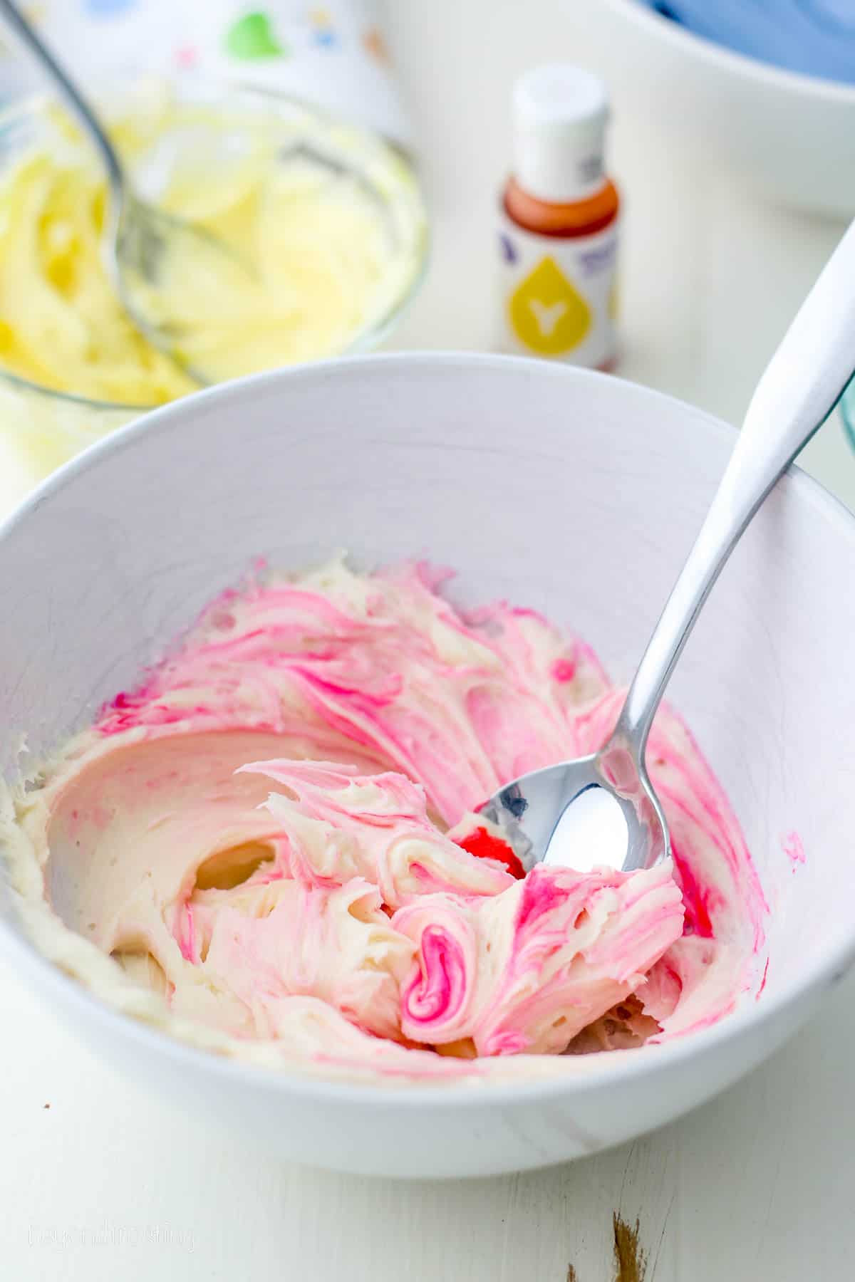 A spoon stirs pink food dye into a bowl of white buttercream frosting.