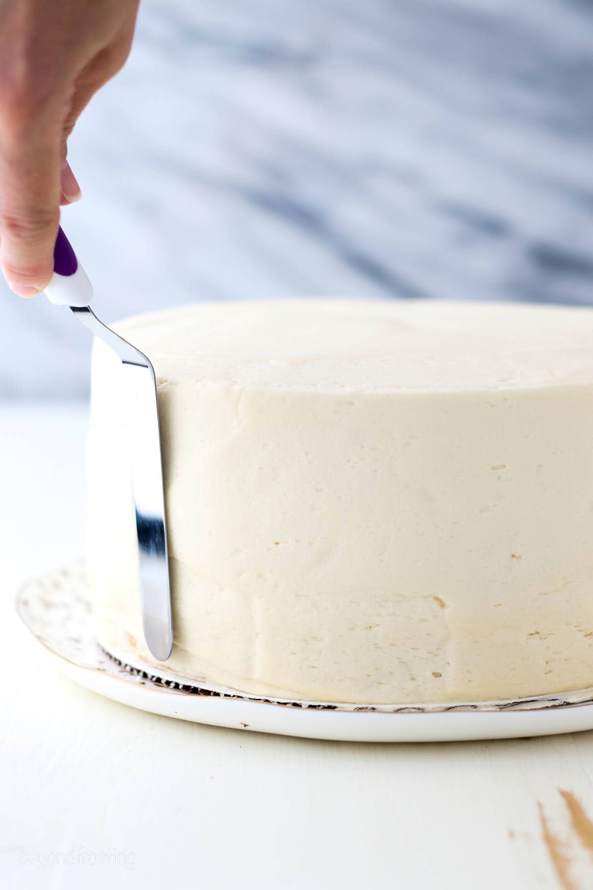A hand using an offset spatula to smooth frosting over the sides of a cake.