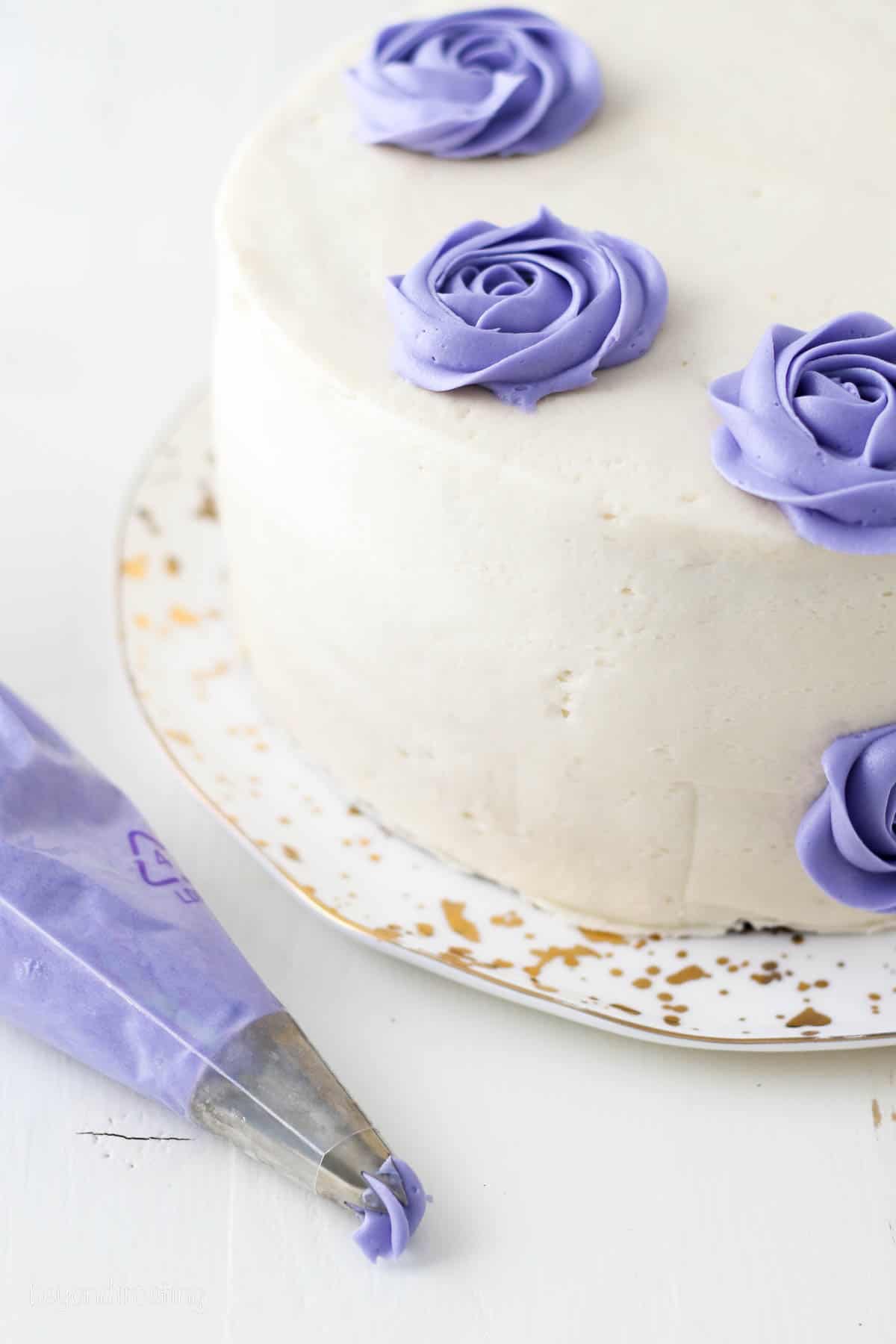 A piping bag filled with purple buttercream next to a cake frosted white with purple buttercream roses.