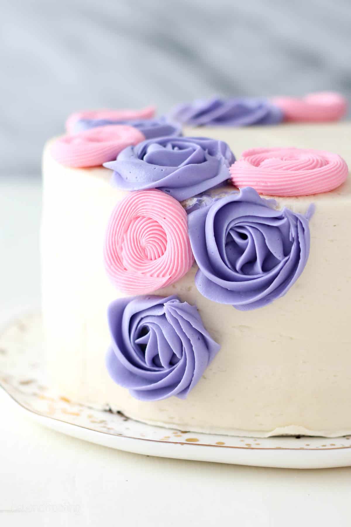 Side view of a cake frosted white, partially decorated with purple and pink buttercream roses.