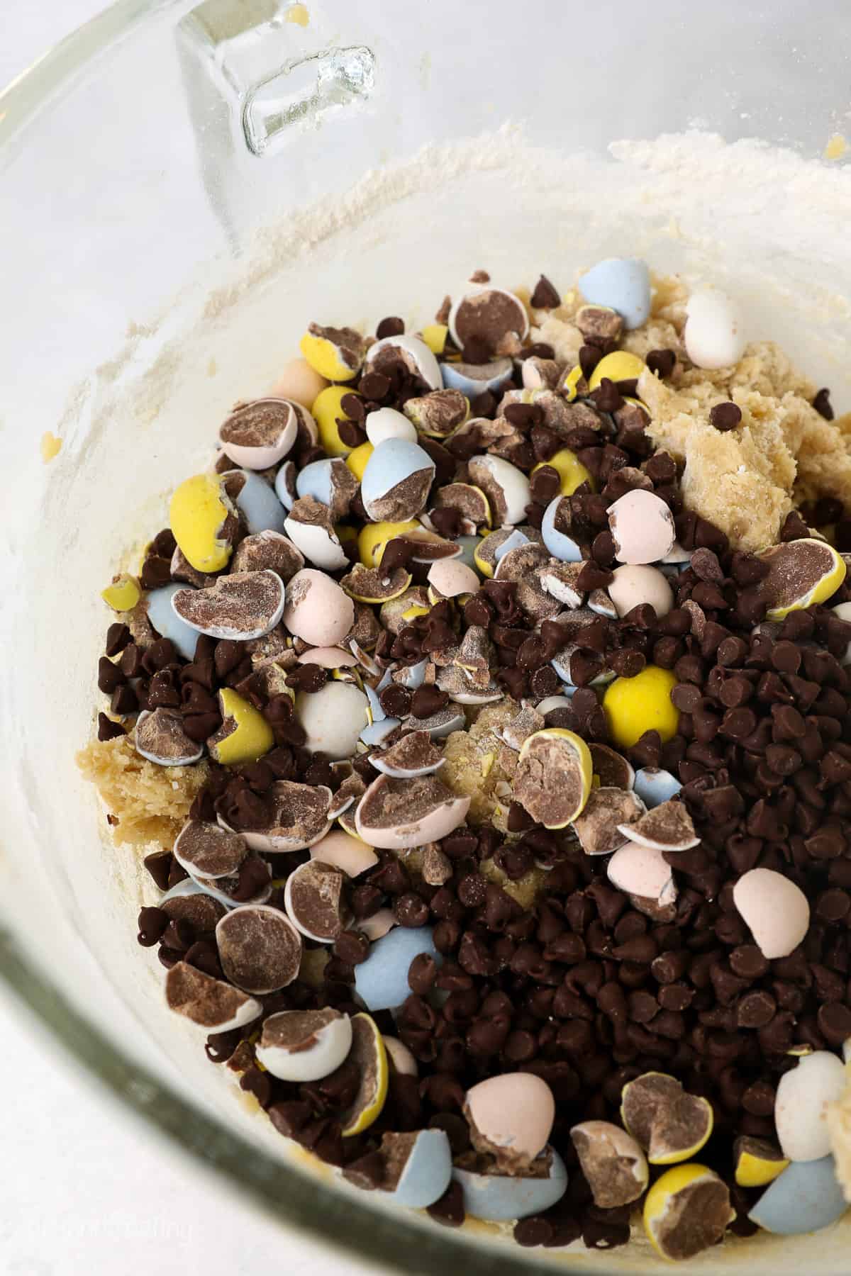 A close up of the inside of a glass mixing bowl showing mini chocolate chips and crushed Cadbury Mini Eggs