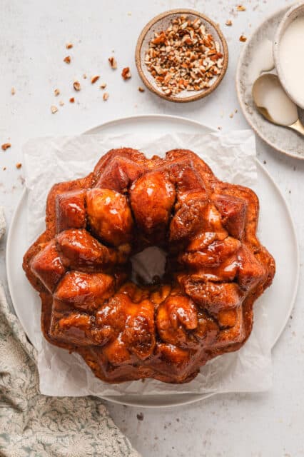 Overhead view of monkey bread turned out on a white plate, next to bowls of vanilla glaze and pecans.