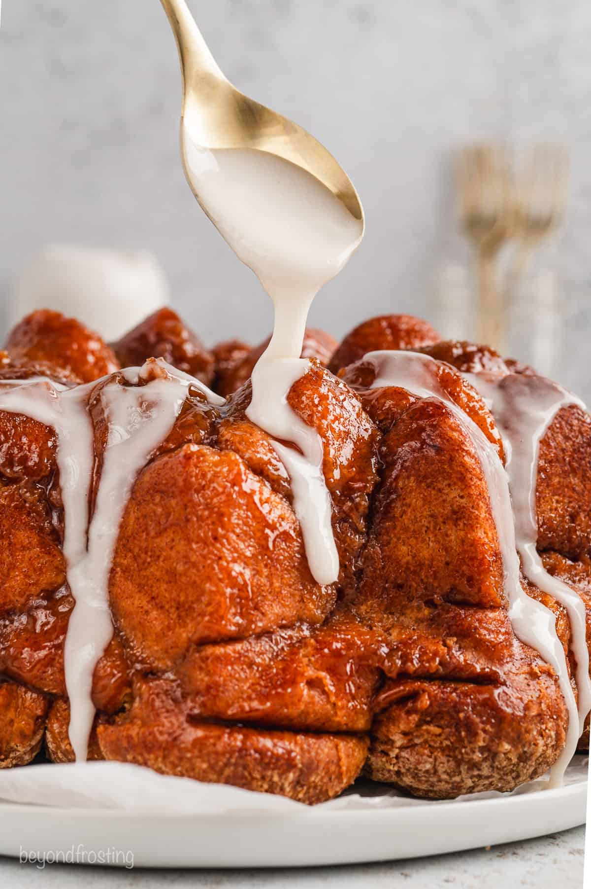 A spoon drizzling vanilla glaze over monkey bread on a white plate.