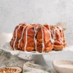 Side view of monkey bread topped with drizzles of vanilla glaze on a cake stand next to bowls of pecans and glaze.