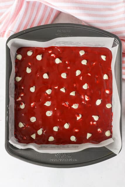 Red velvet brownie batter with white chocolate chips spread into a square baking pan lined with parchment paper.