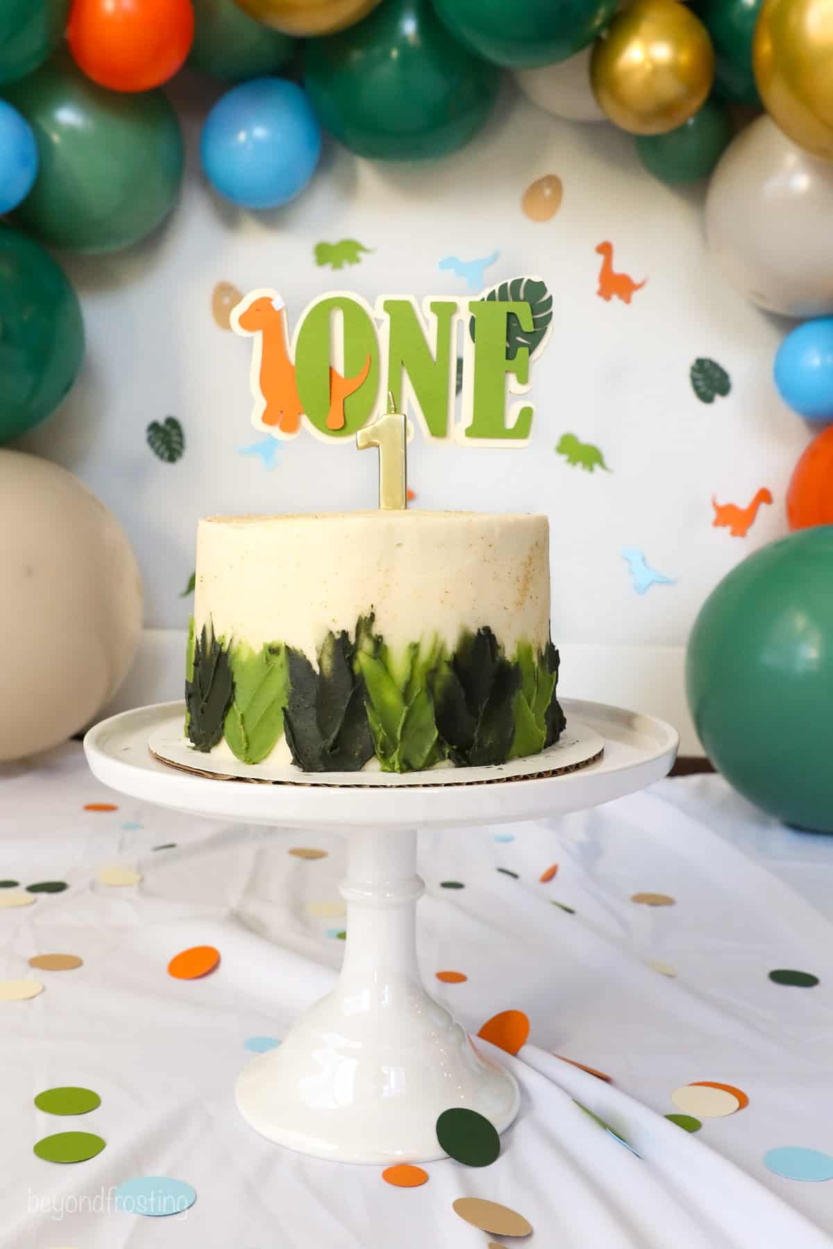 Full shot of a decorated dinosaur cake on a cake stand surrounded by confetti and a birthday hat, framed by a balloon arch in the background.
